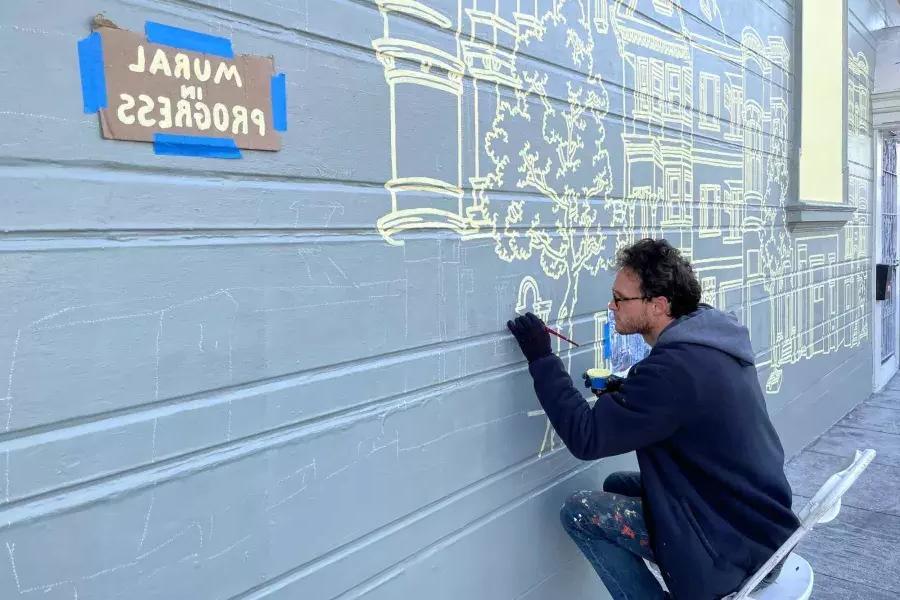 一位艺术家在教会区一栋建筑的侧面画壁画, with a sign taped onto the building that reads "Mural in Progress.加州贝博体彩app.
